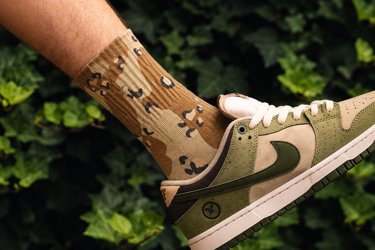 Desert camo printed crew sock worn with an olive and tan sneaker in front of green ivy